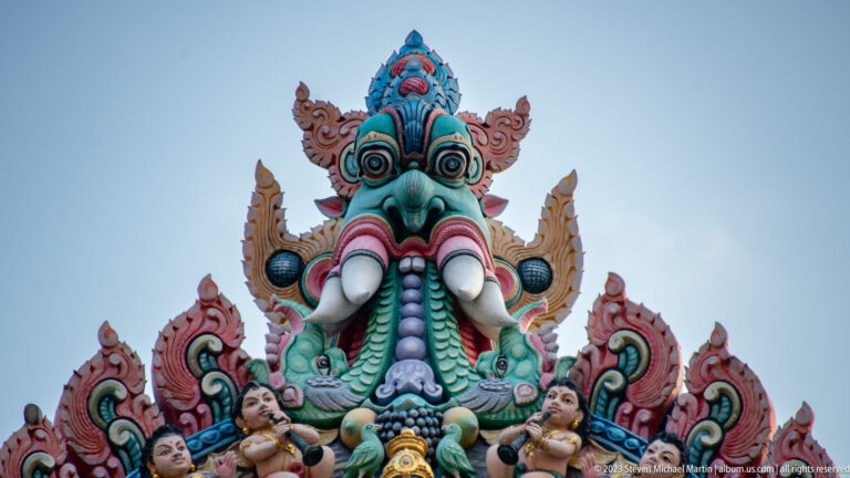 Colorful kirtimukha mask with bulging eyes and tusks at the top of a temple façade.