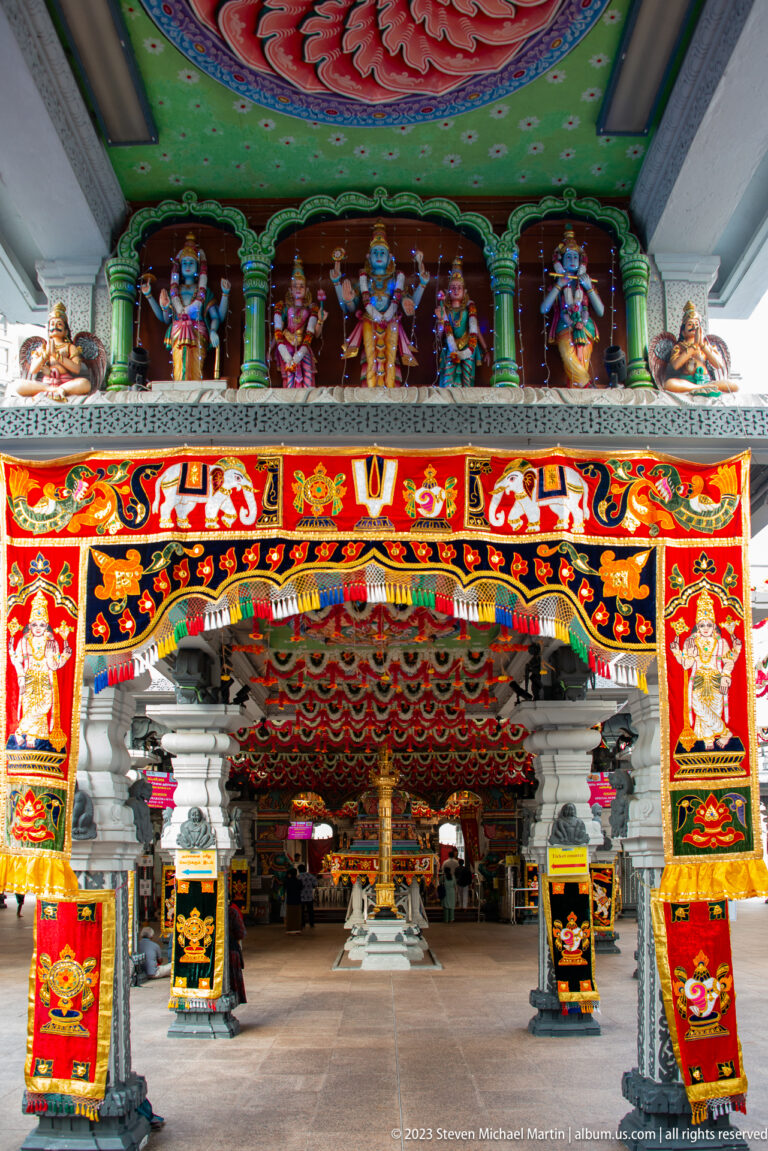 Open temple hall with pale columns, painted ceiling, central pillar, banners, and ritual furnishings.