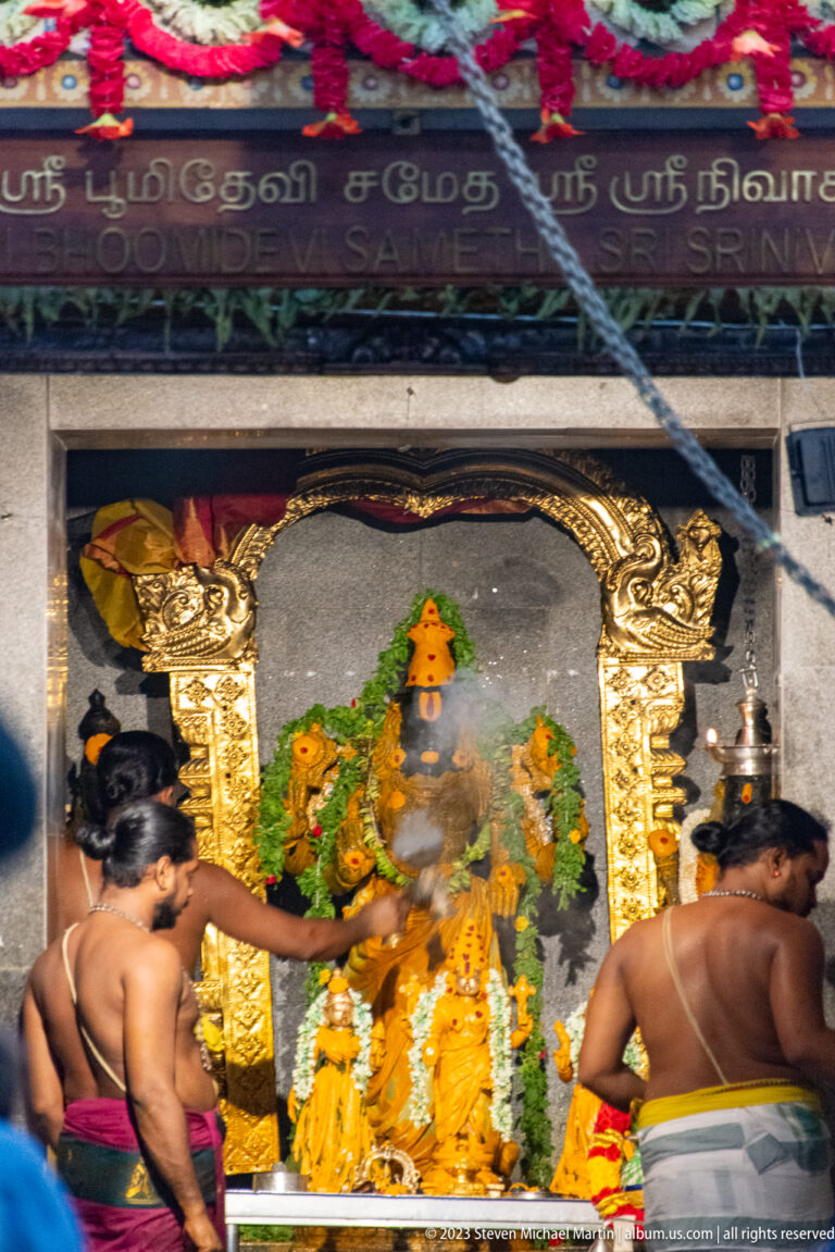 Two Hindu priests performing ritual before a garlanded shrine filled with deity images and offerings.