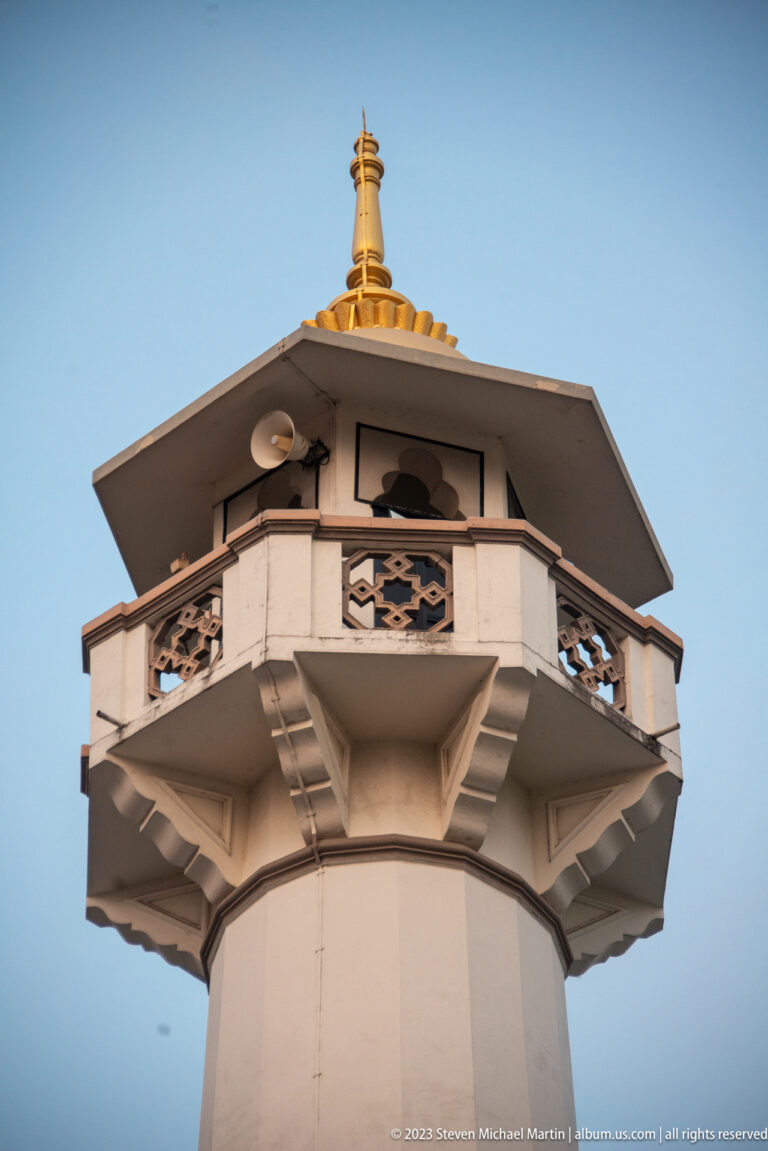 Close upward view of Masjid Sultan minaret with balcony, brackets, openwork panels, and gold finial.