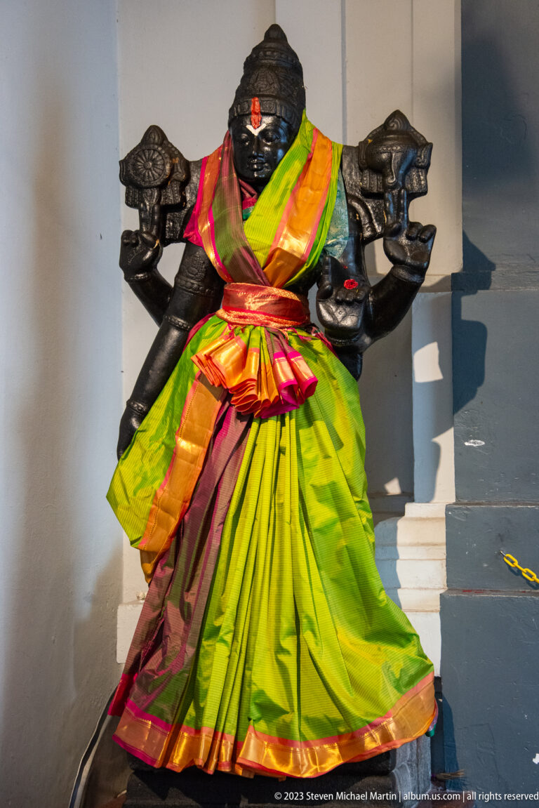 Black stone deity dressed in bright green, magenta, and orange silk with a red forehead mark.