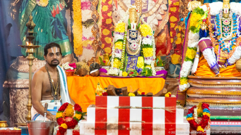 Temple priest seated beside a garlanded shrine with deity images, brass lamp, and striped altar base.