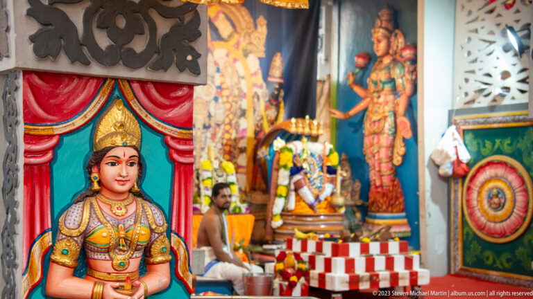 Painted crowned goddess framed by red drapery, with temple shrine and priest blurred behind.
