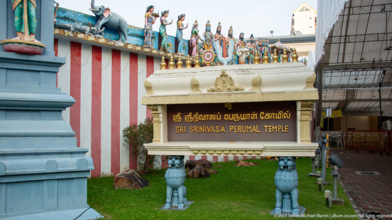 Freestanding Sri Srinivasa Perumal Temple sign beside striped wall and sculpted roofline.