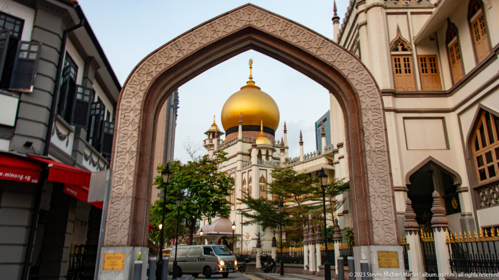 Pointed decorative gateway framing Masjid Sultan’s golden dome, minaret, street, and surrounding buildings.