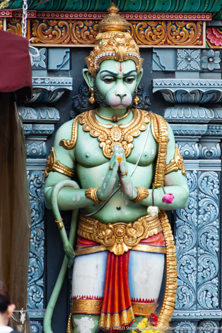 Painted Hanuman with monkey face, long tail, crown, and hands joined in prayer.