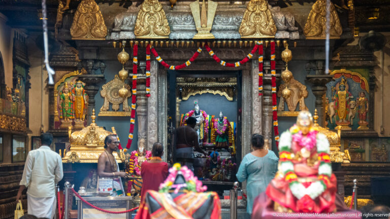 Temple interior with garlanded shrine, carved pillars, hanging decorations, and worshippers.