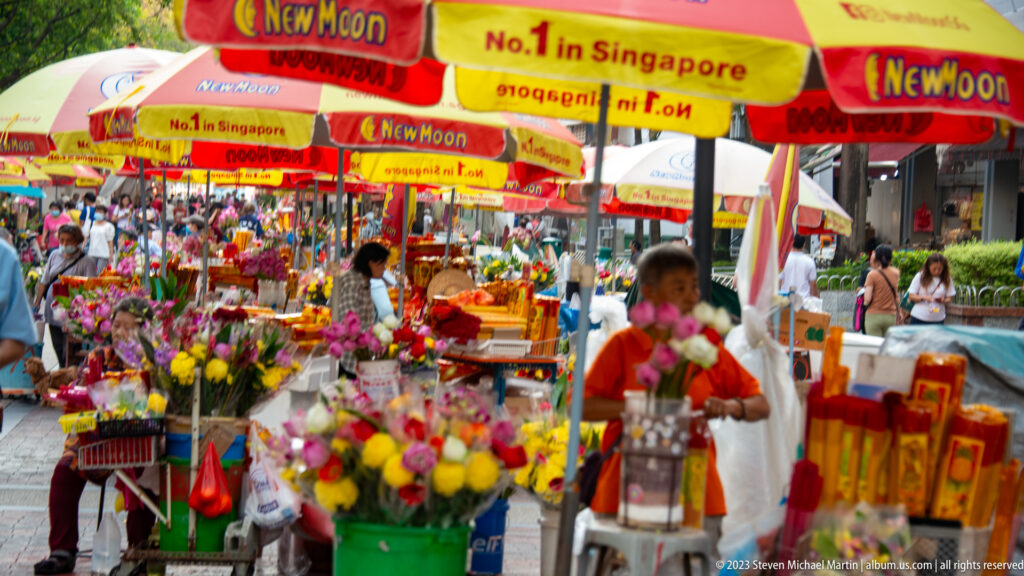 Busy outdoor flower market with bouquets, vendors, customers, and red-yellow umbrellas.