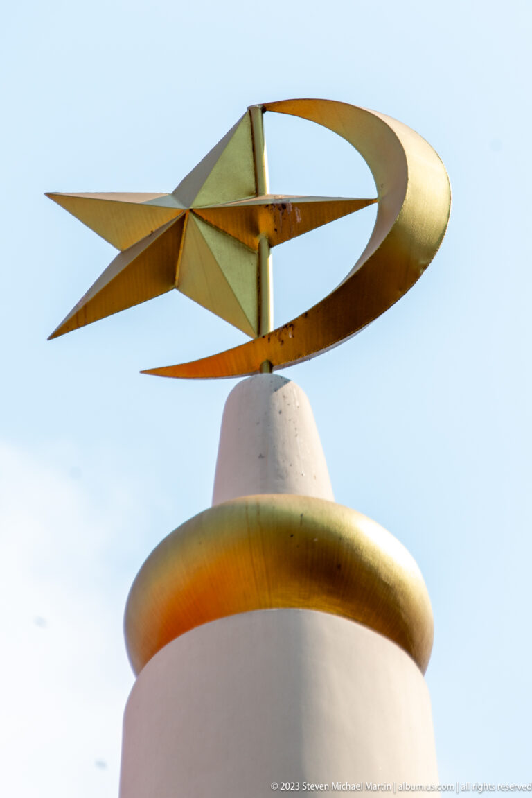 Golden crescent-and-star finial mounted above a white dome against pale blue sky.