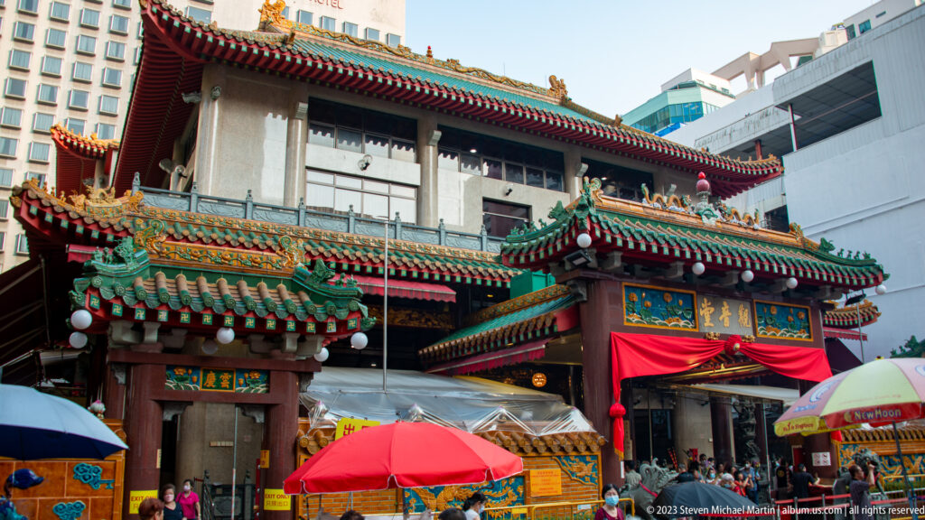 Street entrance of an ornate Chinese temple with layered roofs, painted brackets, umbrellas, and modern buildings behind.