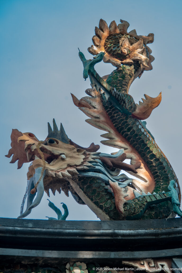 Weathered dragon-like ceramic roof figures with fins and open jaws silhouetted against blue sky.