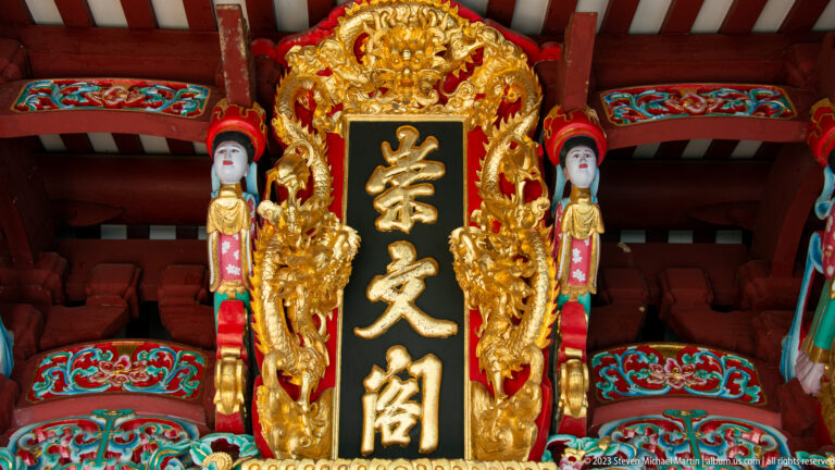 Vertical black temple plaque with gold Chinese characters, carved dragons, and small attendant figures.