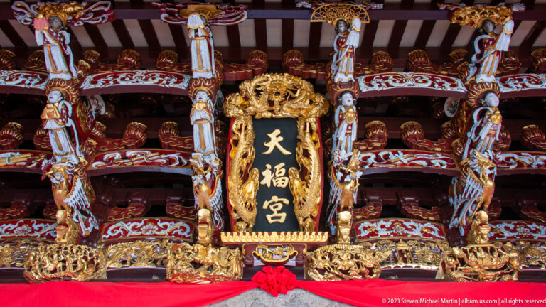 Black-and-gold Tian Fu Gong plaque beneath carved dragons, birds, painted brackets, and red drapery.