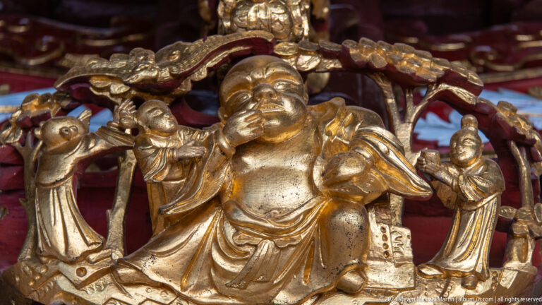 Gilded Laughing Buddha seated beneath an arch with several smaller attendant figures.