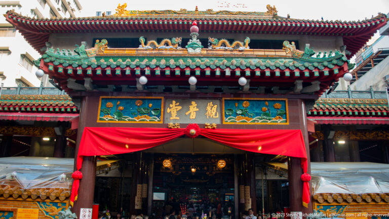 Frontal Chinese temple entrance with green-tiled eaves, gold-lettered signboard, red drapery, and hanging lanterns.