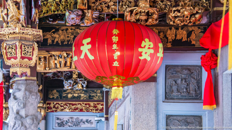 Large red Chinese temple lantern with gold characters hanging beneath carved wooden panels.