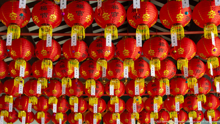 Rows of red prayer lanterns with gold characters and hanging white dedication tags.