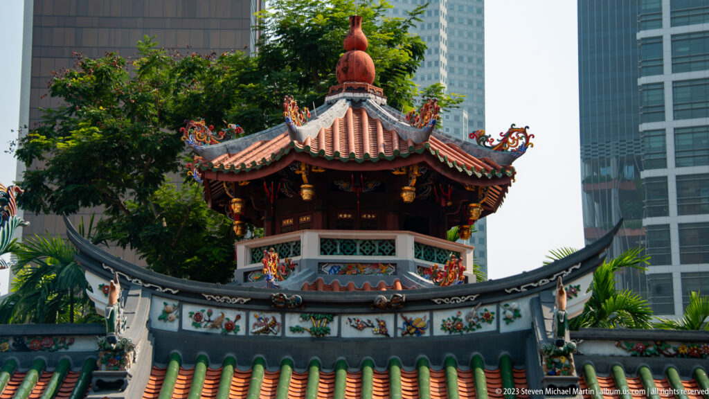 Octagonal Chinese pavilion above a curved green-tiled wall with modern office towers behind.