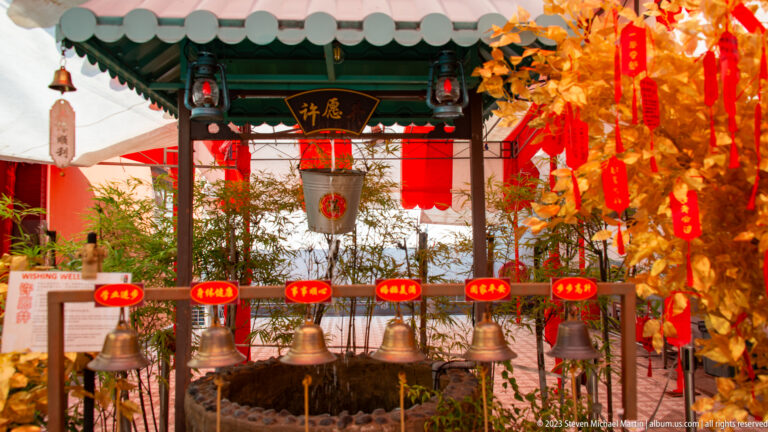 Metal bells beneath a green-roofed pavilion surrounded by red lanterns and gold wish papers.