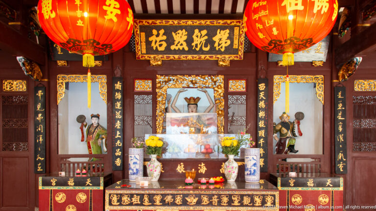 Wooden Chinese temple altar with central sacred image, yellow flowers, offerings, calligraphy, and red lanterns.