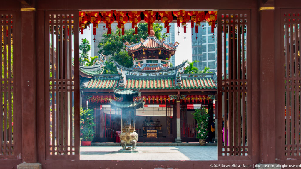 Open timber doors frame a temple courtyard with green roofs, red decorations, pavilion, and incense vessel.