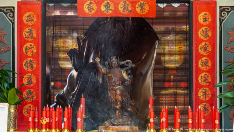 Dark wooden deity figure in a red shrine behind rows of red candles and Chinese inscriptions.