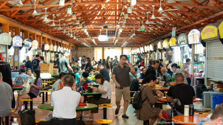 Busy hawker centre with food stalls on both sides, communal tables, diners, and exposed roof trusses.