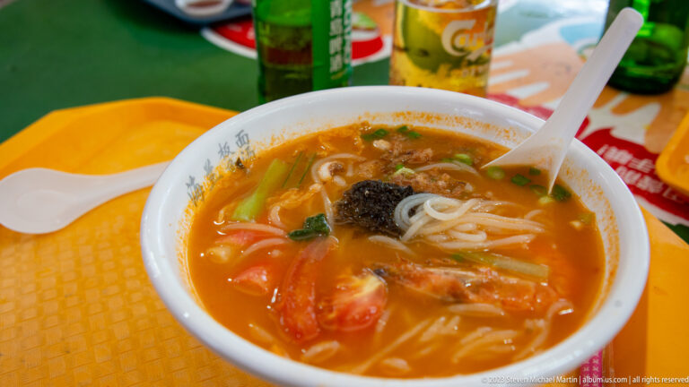 Bowl of orange-red noodle soup with tomatoes, greens, white noodles, and dark garnish on a yellow tray.