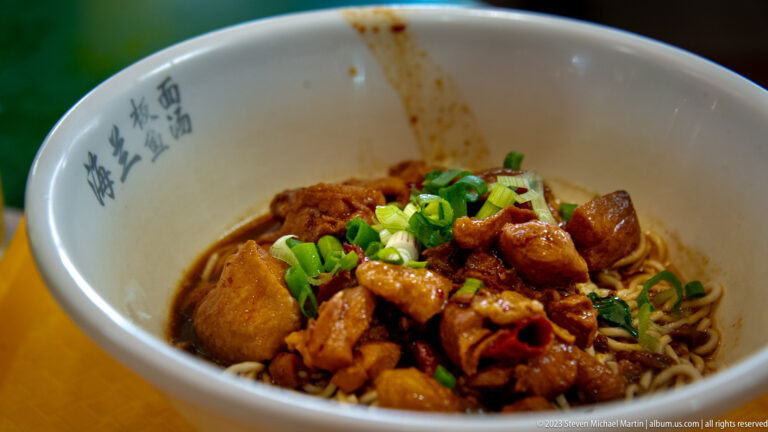White bowl of dry noodles topped with braised meat, sauce, and chopped scallions.