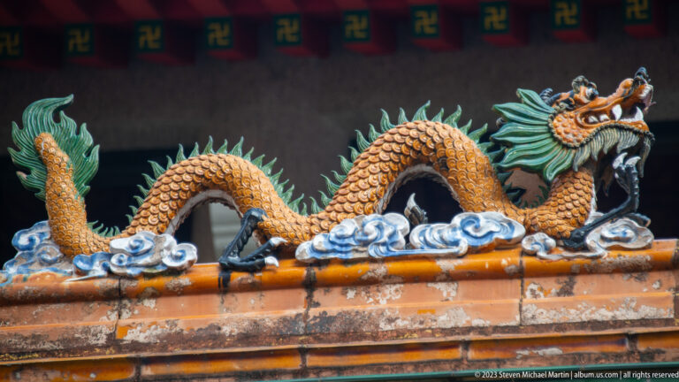 Glazed ceramic dragon with raised claws and open mouth above blue clouds and orange roof tiles.
