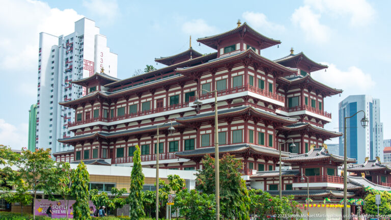 Buddha Tooth Relic Temple exterior with red-and-white galleries, tiered roofs, trees, and modern towers behind.