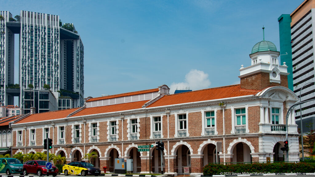 Long colonial-era building with white arcades, red roof, central clock tower, and high-rises behind.