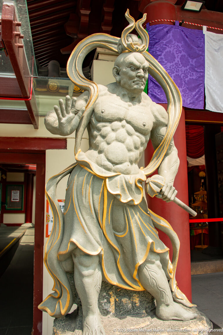 Muscular pale stone temple guardian with flowing drapery and gold-edged details at a red entrance.