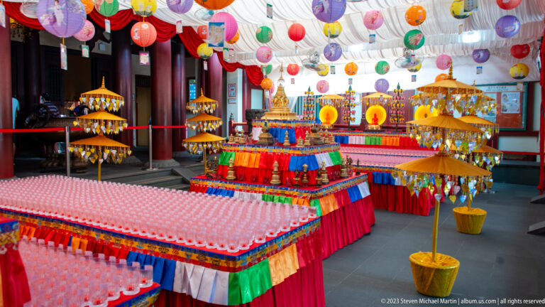 Rows of offering lights and colorful ceremonial objects beneath hanging lanterns in a Buddhist temple hall.