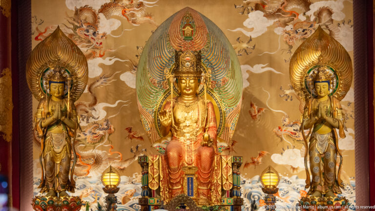 Gilded seated Buddhist figure with large mandorla, flanked by two standing attendants amid clouds and dragons.