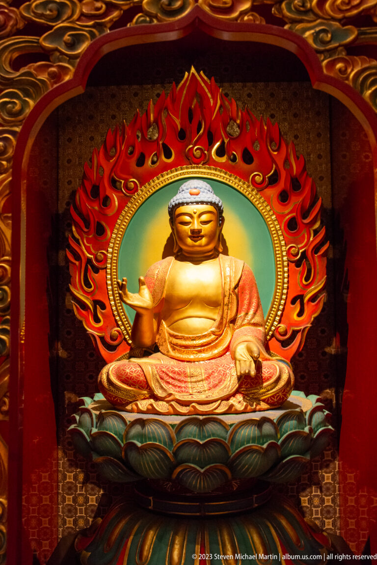 Golden seated Buddha with one lowered hand, lotus throne, circular halo, and red flame mandorla.