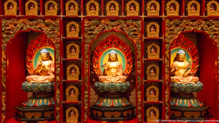 Three seated golden Buddhas in red niches surrounded by rows of miniature Buddha images.