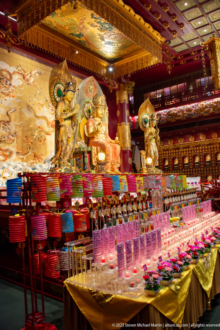 Large gilded Buddhist figures behind offering tables with pink lights, flowers, and colorful papers.