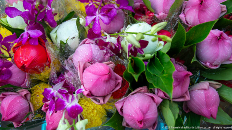 Close bouquet of pink lotus buds, purple orchids, green leaves, and pale flowers.