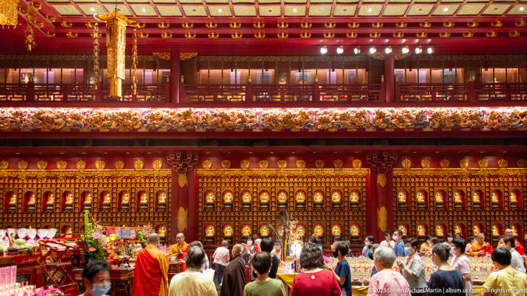 Congregation and monks face a large illuminated wall of Buddha niches in a temple hall.