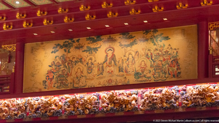 Large Buddhist mural with central haloed figure and many attendants above a carved temple frieze.