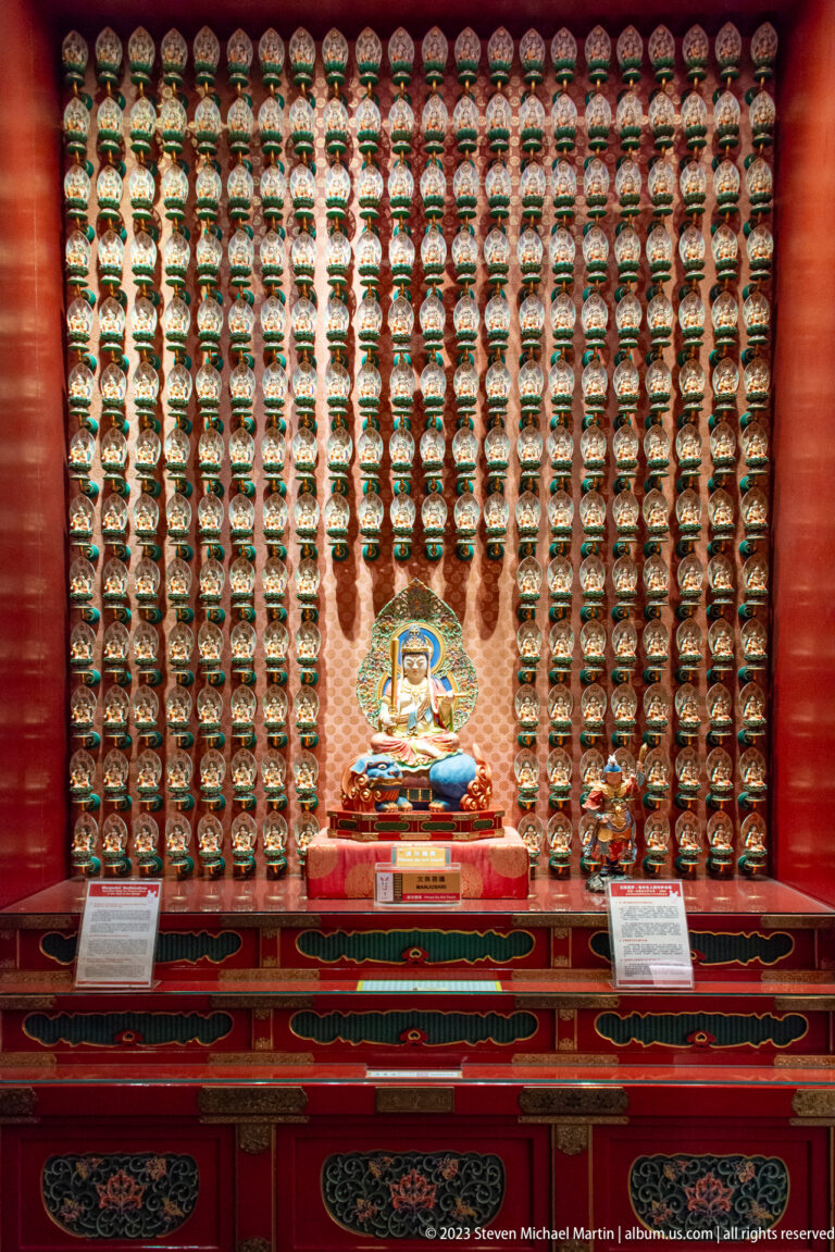 Central seated Buddhist figure before a wall of hundreds of miniature Buddha niches in red cabinetry.