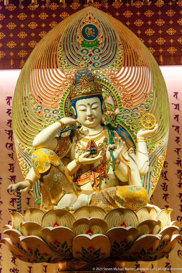 White seated bodhisattva with ritual objects, elaborate crown, lotus base, and multicolored mandorla.