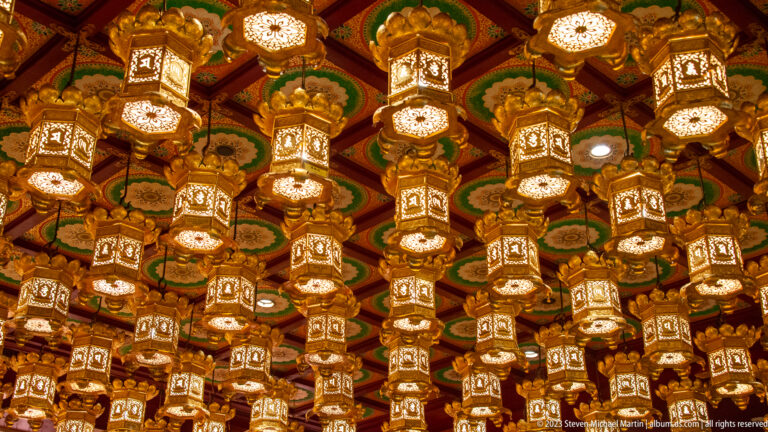 Rows of ornate illuminated lanterns hanging from a richly patterned temple ceiling.