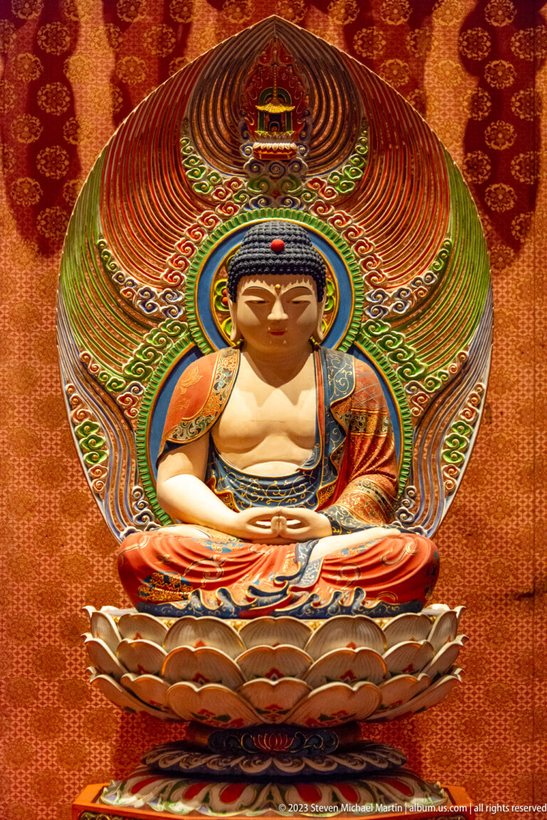Golden seated Buddha in red robe on lotus pedestal beneath a large multicolored mandorla.