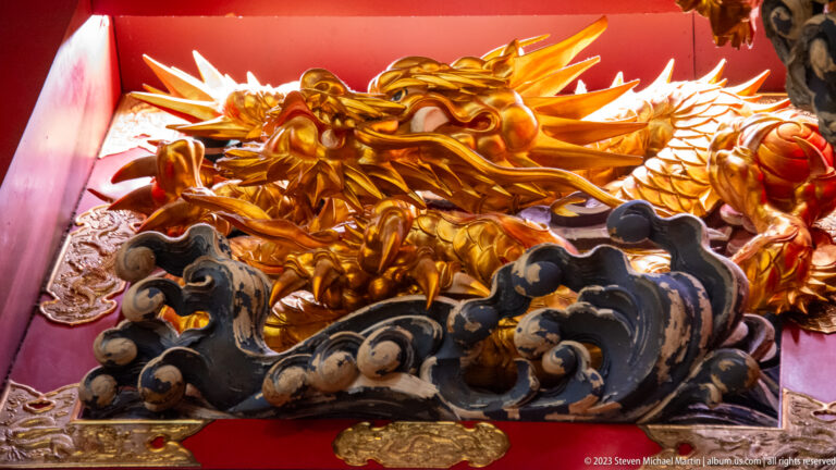 Gilded dragon with open jaws and horns rising above dark sculpted waves against a red background.