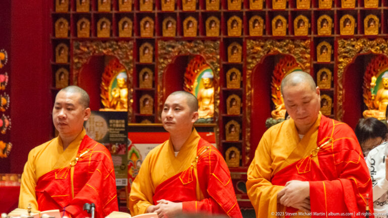 Three Buddhist monks in saffron and red robes standing before illuminated Buddha niches.