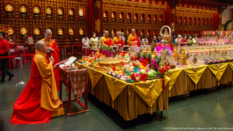 Monks in red and saffron robes beside offering tables filled with fruit, flowers, and ceremonial objects.