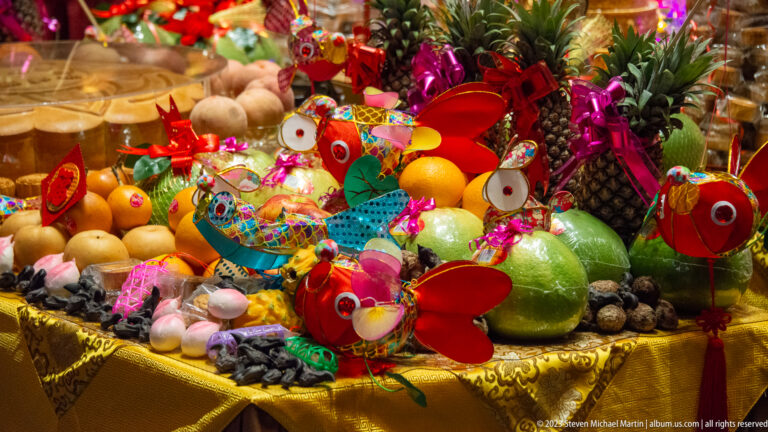 Colorful fruit offerings with pineapples, pomelos, citrus, paper ornaments, and metallic ribbons on gold cloth.