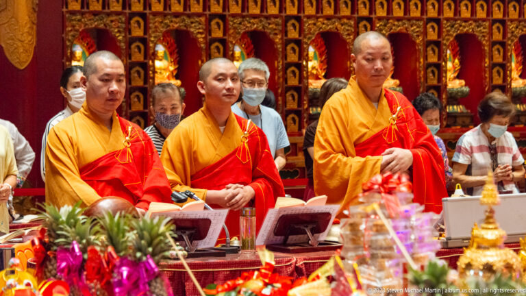 Three Buddhist monks in saffron and red robes standing behind lecterns during a ceremony.